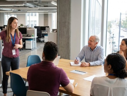 Mahasiswa muda mempresentasikan ide proyek di depan tim, suasana meeting room modern, menambah pengalaman magang nyata.
